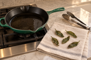 Five crisp fried sage leaves draining on clean linen beside an Artichaut Le Creuset skillet